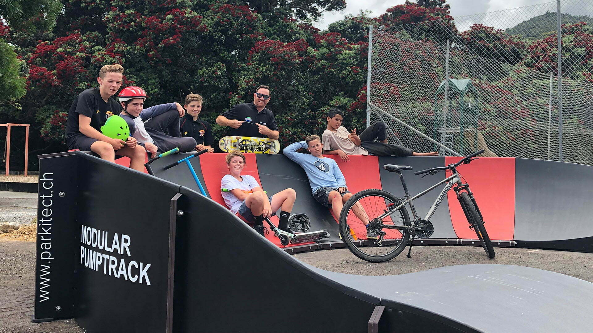 Kids and adults posing on a black and red Parkitect Modular Pumptrack with scooters, skateboards and a bike